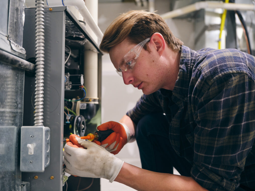 HVAC technician working on residential furnace while wearing white work gloves, safety glasses, and blue plaid button-up shirt.